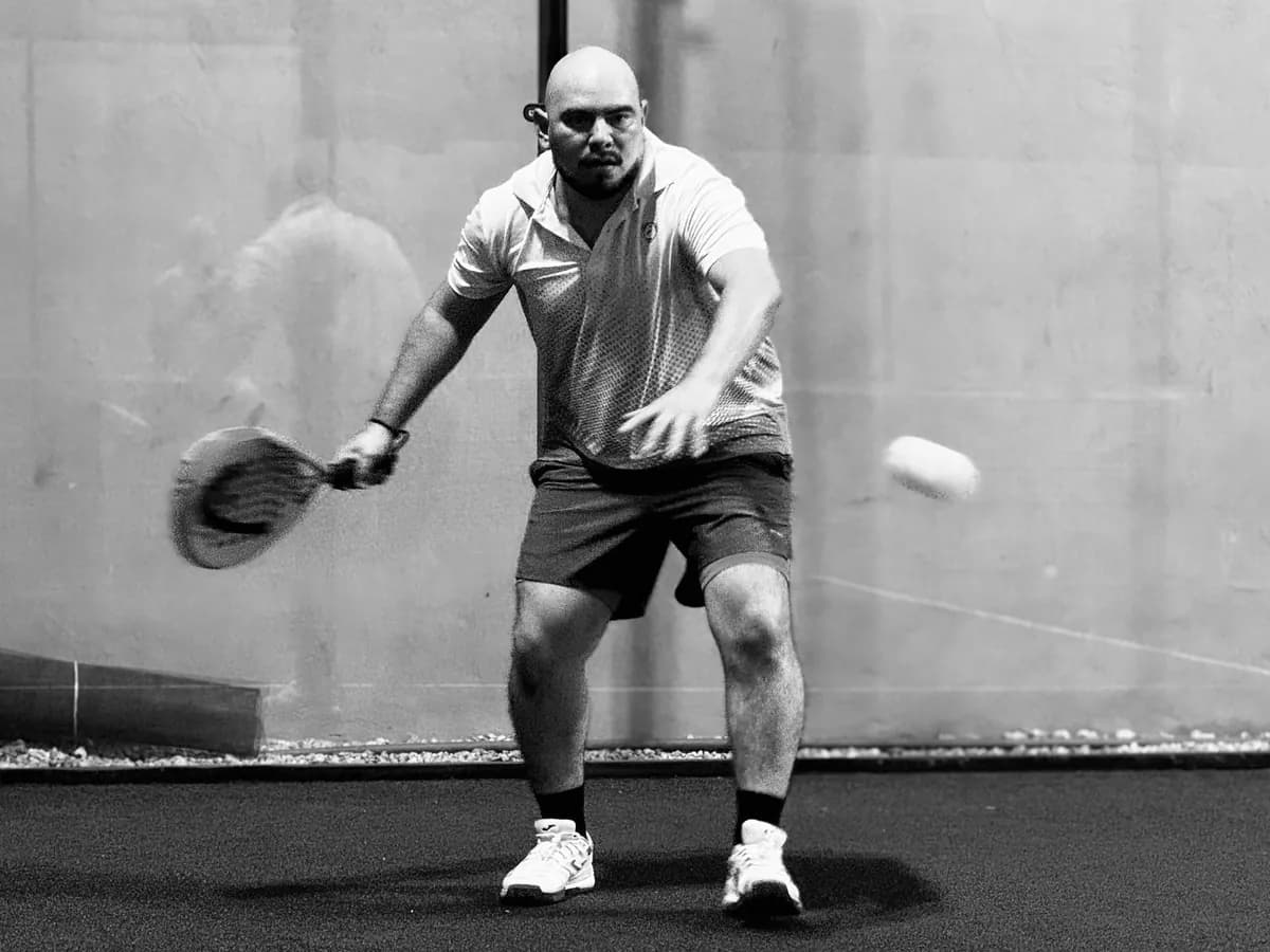Joueur de padel qui frappe un coup droit bas devant la vitre du fond, en noir et blanc