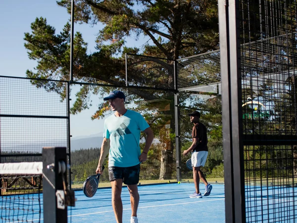 Deux joueurs de padel sur un terrain bleu en plein air, entre le grillage et les vitres